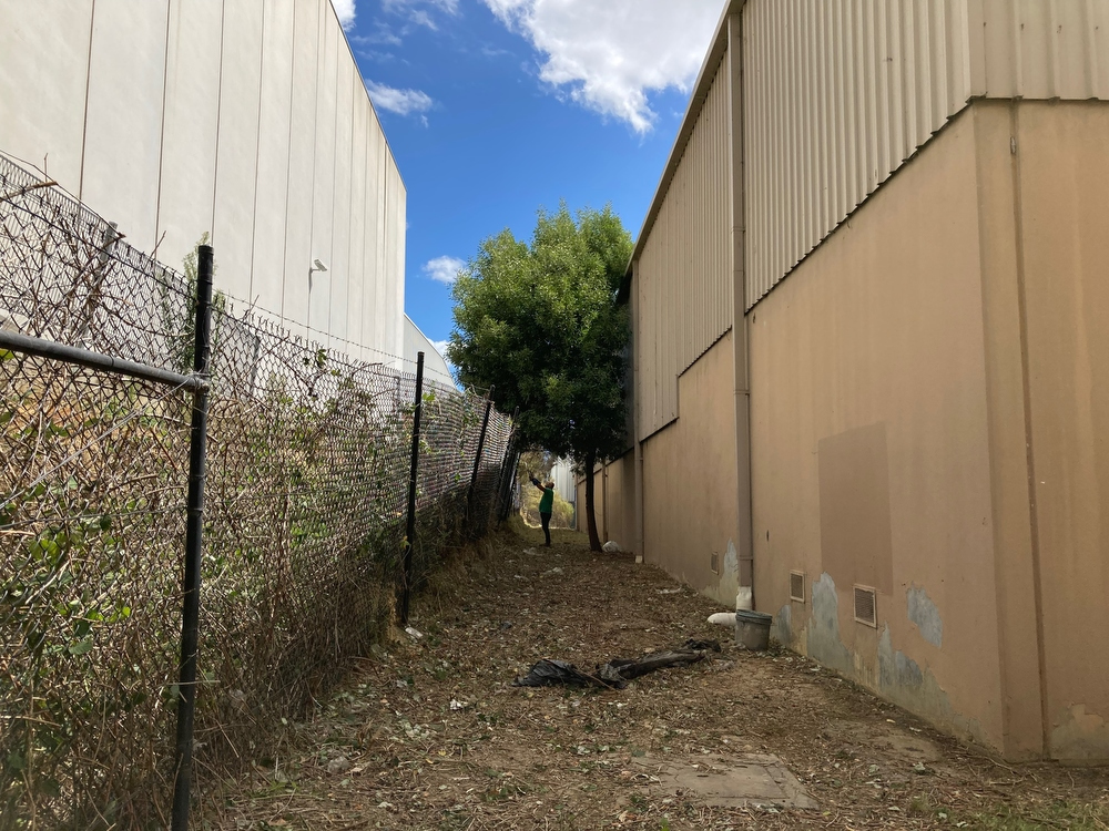 AFTER - Area cleared and debris removed between chain link fence and back wall of industrial building.